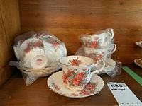 Tea cups and saucers set with floral Centennial Rose pattern, gold trim, some wrapped in plastic bags.