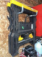 Sawhorse folded flat showing black and yellow plastic frame with metal hooks on sides and supporting yellow plastic pieces inside the fold.
