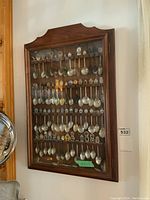 Front view of a solid wood and glass display case containing multiple rows of collectible spoons with decorative handles.