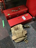 Two red metal tool boxes stacked on a floor next to a beige leather tool belt with pockets and adjustable strap.
