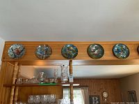 Photo showing five decorative wall plates with colorful bird designs mounted with wire hangers above a doorway or wall shelf.