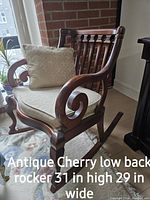 Antique cherry wood low back rocking chair with beige covered seat cushion and pillow shown in room corner near window and carpet.