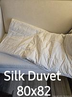 Silk duvet folded and placed on a beige sofa, showing quilted pattern and cream color with dimensions 80x82 inches.