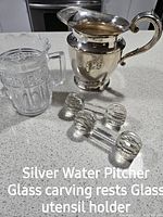 Silver water jug with patina shown alongside glass carving rests and utensil holder on a counter.