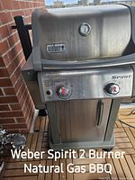 Photo shows a Weber Spirit 2 burner natural gas BBQ with stainless steel lid featuring built-in temperature gauge, two burner control knobs, and cabinet storage below.