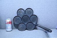 Front view of the cast iron pan showing seven circular cooking sections and a handle, next to a Diet Coke can for size reference.