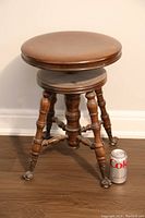 Full view of the antique wooden adjustable piano stool with round cushioned brown seat and turned legs ending in claw metal feet casters, shown next to a soda can for scale.