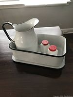 White enamel 8-inch high jug sitting inside a rectangular enamel container along with two milk glass salt and pepper shakers with red lids.