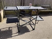 Fully assembled 34 inch high outdoor folding camping table with gray slatted tabletop, two side trays with cup holders, and lower wire shelf rack.