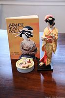 Lot includes a Japanese paper dolls book, a standing geisha doll figurine, and a ceramic wall pocket.