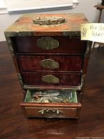 Front view of wooden jewelry box showing four drawers with metal hardware and decorative corner fittings, one drawer open containing small items.