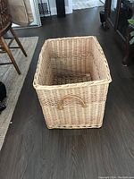 Large rectangular wicker basket with one side handle, shown standing on wooden floor next to furniture and a soda can for scale.