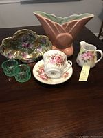Overview of all items on dark wood table: pink scalloped ceramic vase, decorative floral glass dish, two green Vaseline glass holders, ceramic floral pitcher, and Royal Sutherland Bone China cup and saucer