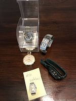 Five watches displayed on a table including pocket watch, ladies wristwatches, Swatch Skin in case, and digital Casio watch