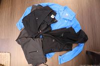 Four outdoor clothing items laid flat on wooden floor, including two pairs of black pants, a blue pullover, and a black hoodie.