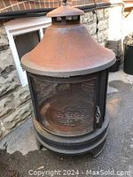Full side view of heavy steel outdoor firepit showing rusted cone-shaped chimney top, mesh door and ventilation base.