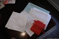 Top down photo showing five folded linens and tablecloths in white, light blue-green, and red color, displayed on dark wood surface.