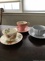 Three bone china cups and their matching saucers displayed on a dark wooden table under a window; pink, blue, and floral patterned cups shown side by side.