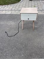 Full view of wooden night stand showing single drawer, tapered legs, and power cord.