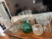 Wide view of the collection showing various clear glass bowls, green glass bottle, crystal cut glass bowl, and vases all on a wooden surface against a dark background