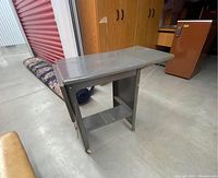 Metal rolling typewriter stand with one side panel extended and one side panel folded, showing industrial grey painted surface and four wheels on bottom.