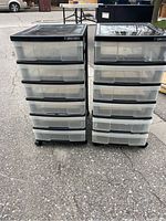 Front view of two plastic storage units with six clear drawers each, black tops and bases on wheels, standing outside on asphalt.