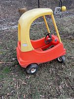 Full profile side view of red and yellow plastic Cozy Coupe toy car outdoors on grass showing body, wheels, and roof.