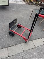 Full side view of red and black metal dolly with adjustable handle and two wheels on pavement.