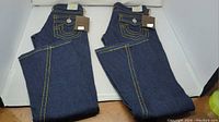 Two pairs of folded dark blue True Religion women's jeans with yellow stitching and tags visible, shown from front side