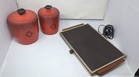 Photo showing two vintage reddish-orange metal canisters with black lids and black decorative floral design around the labeled text alongside Salton Hotray rectangular electric food warmer with ridged surface and side wooden handles.
