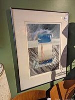 Framed abstract print showing full artwork including mat and frame on a green wall with mixed shapes and colors featuring blues, grays, and red-orange lines.