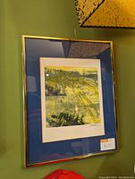 Photo of the framed abstract print featuring green, yellow, and blue colors in a gold frame hanging on a green wall.