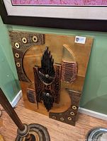 Front view of large rectangular metal art piece positioned on floor against wall, showing abstract geometric design with textured, ribbed and circular metal elements in brass and bronze tones.