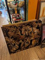 Photo of a large rectangular abstract painting on canvas leaning against a wall. The painting has a brown and yellow background with bold black abstract shapes and lines.