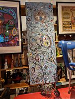 Long vertical abstract painting with colorful swirling shapes and black looping lines on textured light blue background, displayed among other framed art and artifacts.