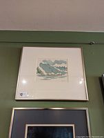 Framed small landscape art piece against green wall with artist signature visible in the lower right corner.