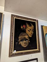 Framed velvet painting showing close-up of two faces on dark background, one adult female with red flower in hair and one child.