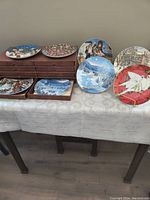 Full view of the set of eight decorative plates placed on a table, some on top of a wooden storage box with pull-out drawers, and others standing upright beside it.