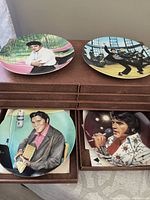 Top view of four Elvis Presley plates placed on a brown storage box with drawers: one with pink car background, another with a dancing prison stripes theme, one holding a jukebox microphone, and another singing with a white jumpsuit.