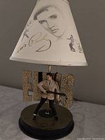 Close-up photo of lamp showing detailed Elvis Presley figurine playing guitar, glitter gold 'ELVIS' letters, and lampshade with Elvis images and signature.