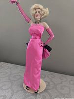 Front view of Marilyn Monroe doll in pink gown and gloves on stand, showing facial features and outfit details.
