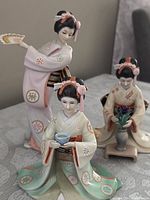 Three Franklin porcelain Asian maiden figurines displayed together on a table: one holding a fan, two holding teacups, all in traditional kimonos with delicate pastel colors and detailed painting.