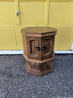 Front view of 1970s octagon wooden side table with storage doors and metal handles against a yellow garage door backdrop.