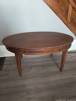 Oval wood coffee table with scalloped edge, drawer on front, and detailed legs, shown from side angle.