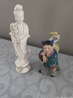 Two porcelain figurines on a patterned surface, showing a white standing woman figure and a colorful girl with geese on her back.