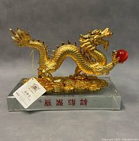 Front angled view of golden dragon figurine on clear glass base with red Chinese characters and gold coin details. Shows dragon holding red orb in claw.