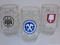 Front view of three dimpled glass beer mugs with German brewery logos, placed side by side.