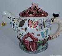 Front angle of white ceramic butterfly teapot candle holder showing painted colorful butterflies, cut-out windows, and thatched roof lid.