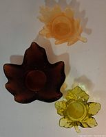 Three maple leaf-shaped glass votive candle holders in amber and brown tones arranged on white surface, shown from above.