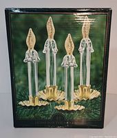 Original packaging box showing the four glass candle clips with gold tone bases and clear glass stems with gold flame tips against a green and black background with holiday theme.